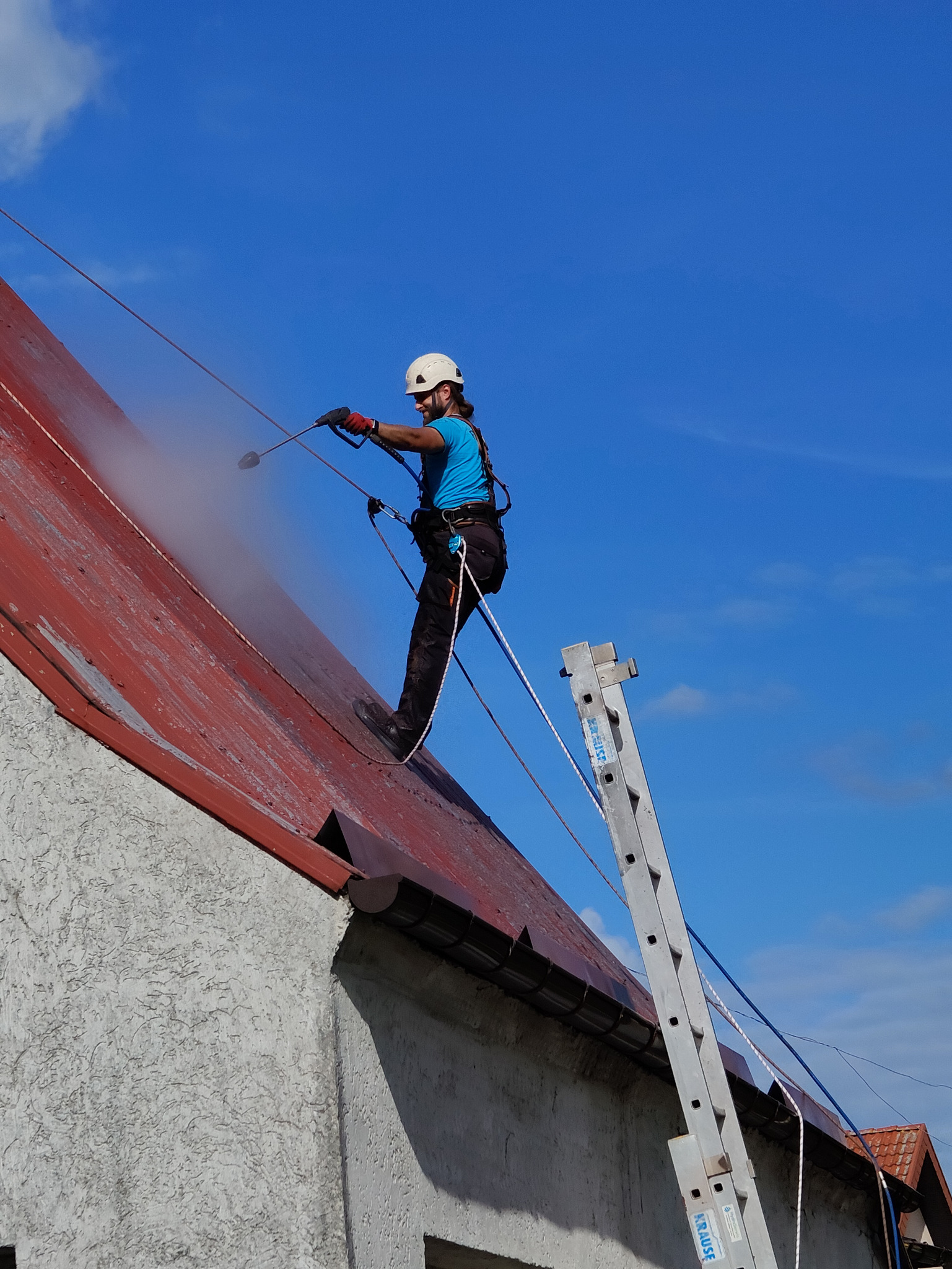 Pracownik w kasku i uprzęży zabezpieczającej czyści czerwoną, metalową dachówkę myjką ciśnieniową, z boku drabina oparta o budynek.