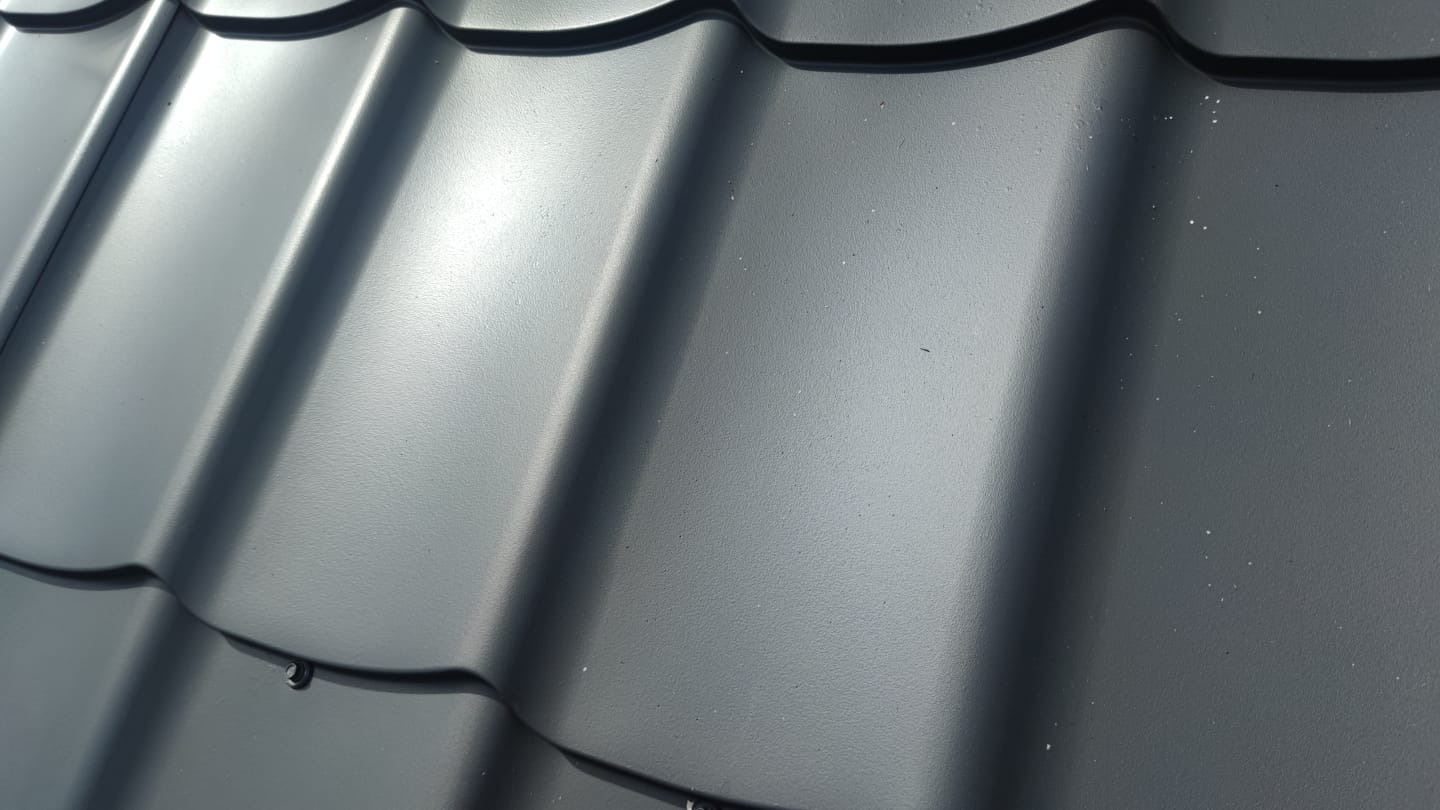 Szczegółowe ujęcie grafitowej blachodachówki z widocznymi śladami po deszczu, ukazujące regularne fale i mocowania. Tekstura powierzchni podkreślona przez oświetlenie.