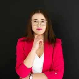 Portret kobiety w różowym żakiecie i okularach, z ręką pod brodą, na czarnym tle.