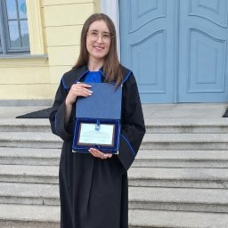 Kancelaria Radcy Prawnego Radca Prawny Weronika Bagrowska - Uśmiechnięta kobieta w todze z niebieskimi wykończeniami, prezentuje dyplom w otwartej niebieskiej kasetce, stojąc przed żółtym budynkiem z błękitnymi drzwiami.