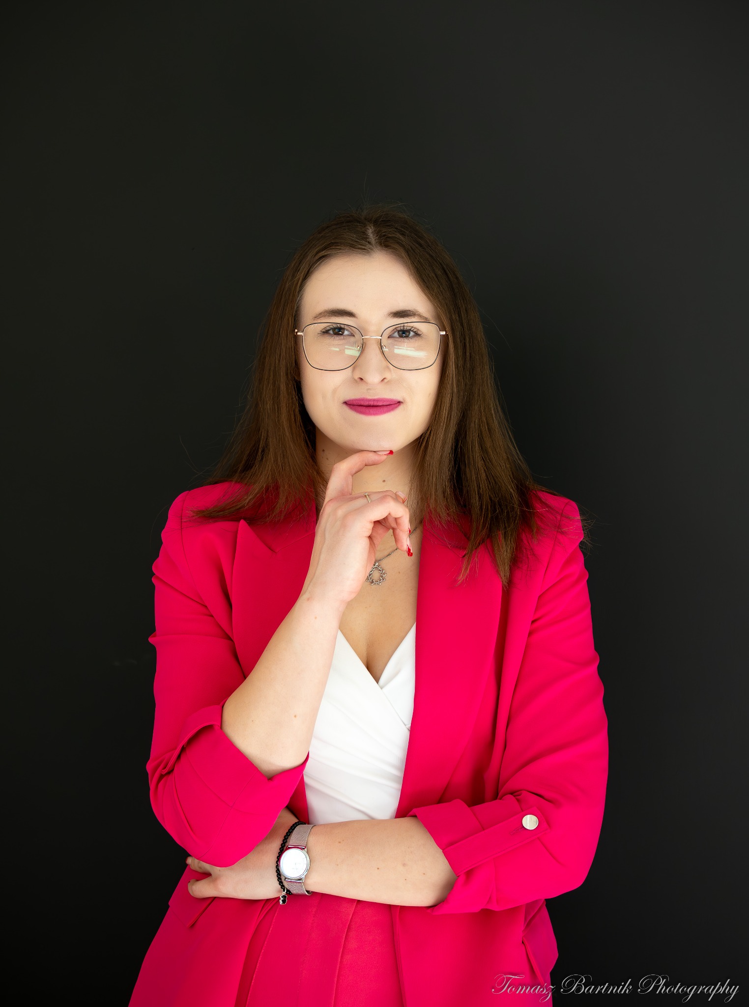 Portret kobiety w różowym żakiecie i okularach, z ręką pod brodą, na czarnym tle.