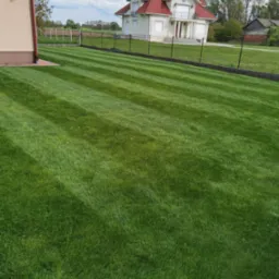 Perfekcyjnie przystrzyżony trawnik z wyraźnymi pasami, otoczony ogrodzeniem z siatki, z widokiem na duży dom z czerwonym dachem i balkonem.