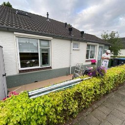 HUBERT NOWAK - Pracownik w białym kombinezonie stoi przed pomalowanym na biało domem z cegły, sprawdzając coś na telefonie. Widoczne okna z kwiatami, drabina oparta o ścianę, zabezpieczona folią podstawa budynku,...