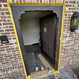 HUBERT NOWAK - Renowacja drewnianych drzwi wejściowych z użyciem taśmy malarskiej i uszczelniacza, widoczne narzędzia i materiały budowlane na zewnątrz.