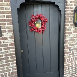 HUBERT NOWAK - Ciemnoszare, świeżo pomalowane drzwi wejściowe z dekoracyjnym wieńcem z czerwonych koralików, zawieszonym na tle ceglanej ściany.