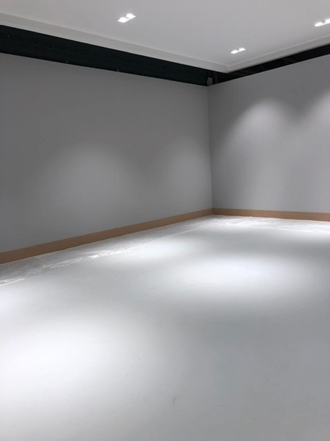 Pusty, świeżo pomalowany pokój z szarymi ścianami i białym podłożem, listwa przypodłogowa w naturalnym odcieniu drewna, oświetlenie sufitowe punktowe.