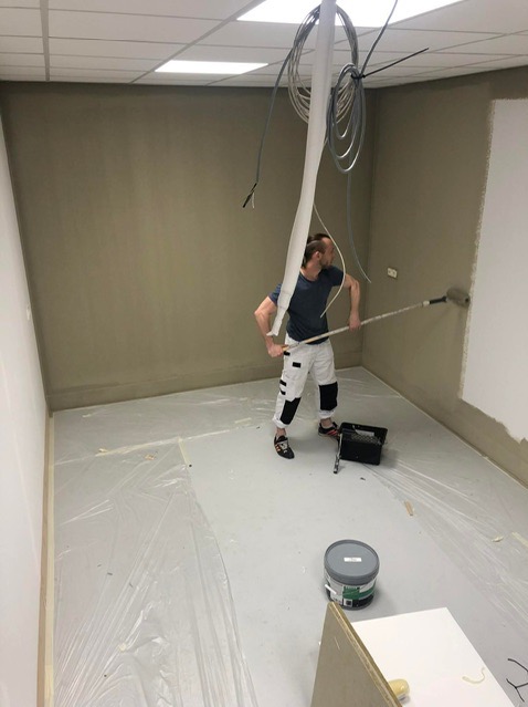 Mężczyzna maluje ścianę na biało za pomocą wałka w pomieszczeniu biurowym z zabezpieczoną folią podłogą, widoczne kable zwisające z sufitu.