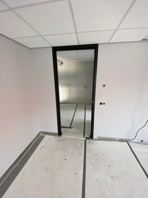 Wnętrze biura w stanie deweloperskim, widoczne białe ściany, sufit kasetonowy, czarna futryna drzwi i podłoga z widocznymi liniami podziału, przygotowana do wykończenia.