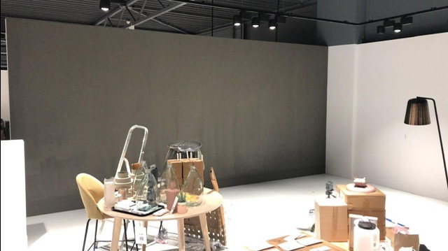 Przygotowania do malowania ściany w odcieniach szarości w przestronnym wnętrzu, widoczne narzędzia i akcesoria malarskie na okrągłym stoliku oraz drabina, lampa podłogowa w rogu pomieszczenia.