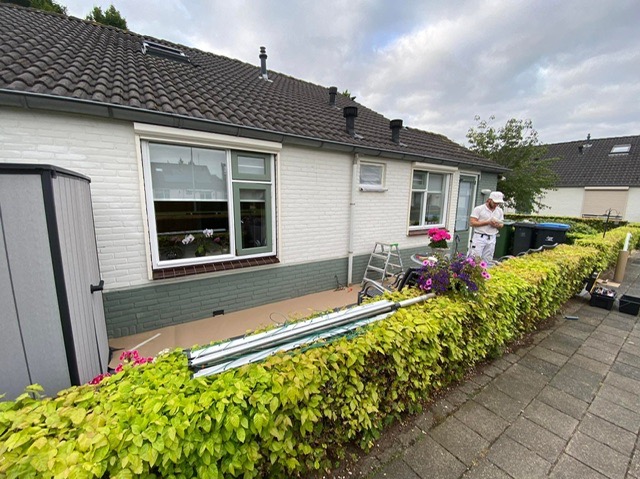 Pracownik w białym kombinezonie stoi przed pomalowanym na biało domem z cegły, sprawdzając coś na telefonie. Widoczne okna z kwiatami, drabina oparta o ścianę, zabezpieczona folią podstawa budynku,...