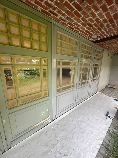 Fragment ściany z trzema segmentami okiennic w trakcie renowacji, z widoczną warstwą starej farby i świeżo nałożonym podkładem, zabezpieczone folią malarską na podłożu, ceglany sufit