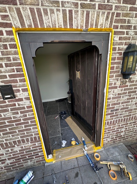 Renowacja drewnianych drzwi wejściowych z użyciem taśmy malarskiej i uszczelniacza, widoczne narzędzia i materiały budowlane na zewnątrz.