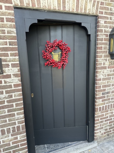 Ciemnoszare, świeżo pomalowane drzwi wejściowe z dekoracyjnym wieńcem z czerwonych koralików, zawieszonym na tle ceglanej ściany.