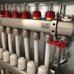 Rozdzielacz instalacji centralnego ogrzewania z rotametrami, z widocznymi białymi rurami i zaworami, zamontowany w szarej szafce.