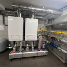 Dwa białe kotły grzewcze Brötje, połączone rurami z instalacją. Widoczne filtry, zawory i manometry. Na ścianie schemat instalacji.