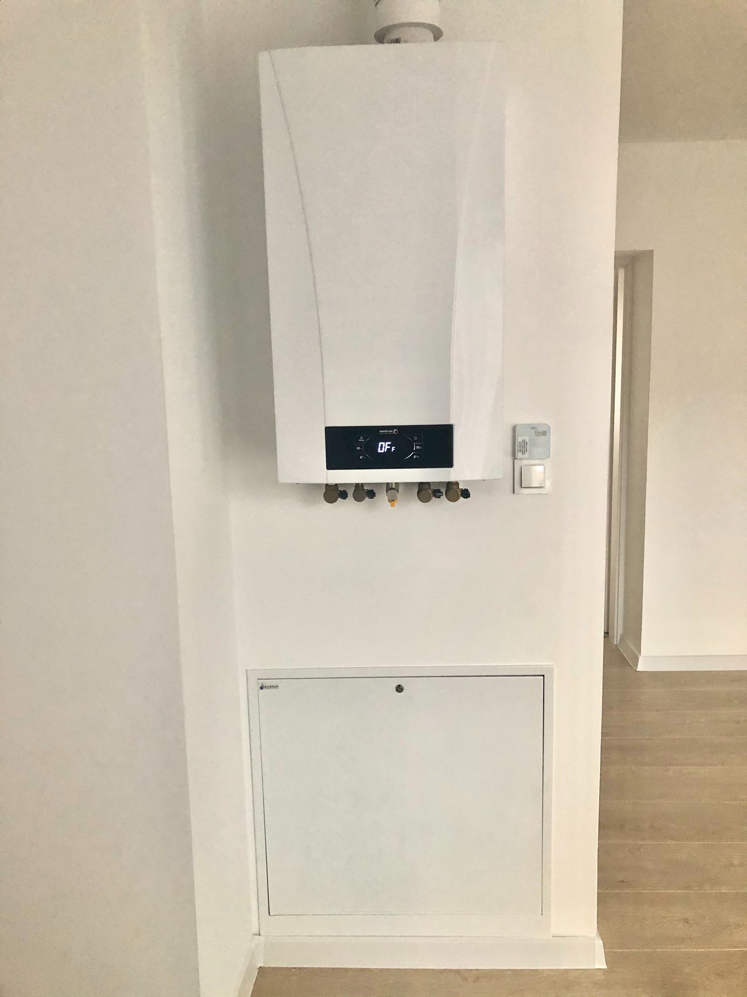 Nowoczesny, biały piec gazowy zamontowany na białej ścianie, widoczny panel sterowania w trybie OFF, zawory przyłączeniowe oraz klapka rewizyjna poniżej, w tle fragment parkietu i drzwi.