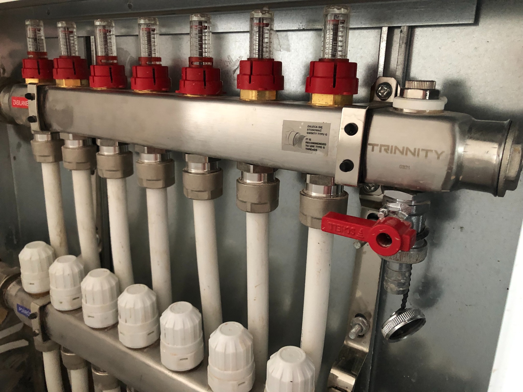 Rozdzielacz instalacji centralnego ogrzewania z rotametrami, z widocznymi białymi rurami i zaworami, zamontowany w szarej szafce.