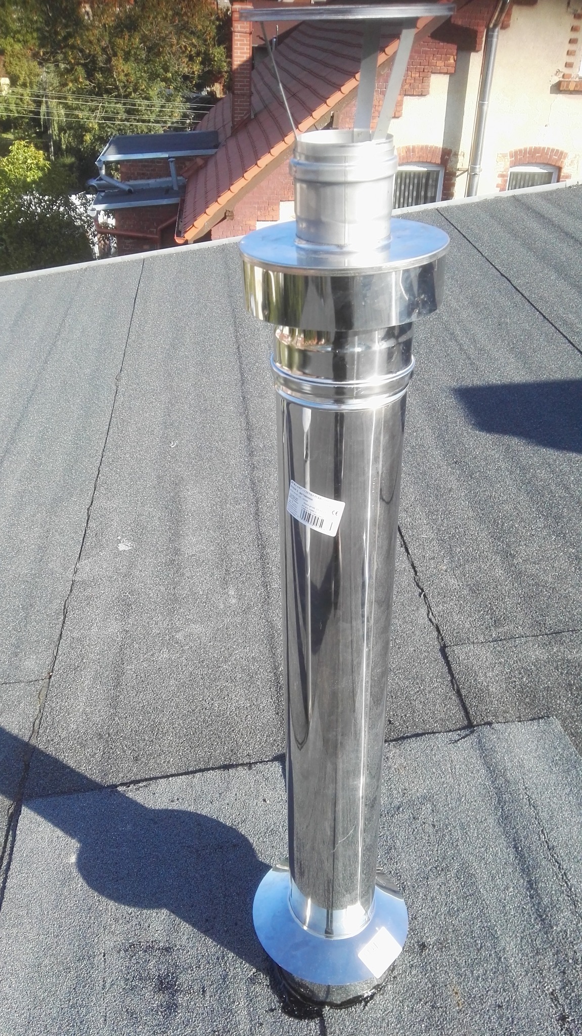 Błyszczący, nowy komin ze stali nierdzewnej zamontowany na płaskim, bitumicznym dachu, z widocznym cieniem i fragmentem budynku w tle.