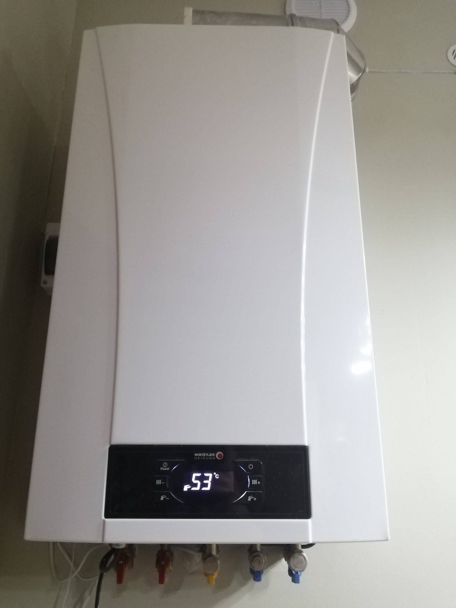 Biały, wiszący piec gazowy marki Brötje z wyświetlaczem wskazującym temperaturę 53 stopnie Celsjusza, zamontowany na jasnej ścianie, z widocznymi przyłączami hydraulicznymi.