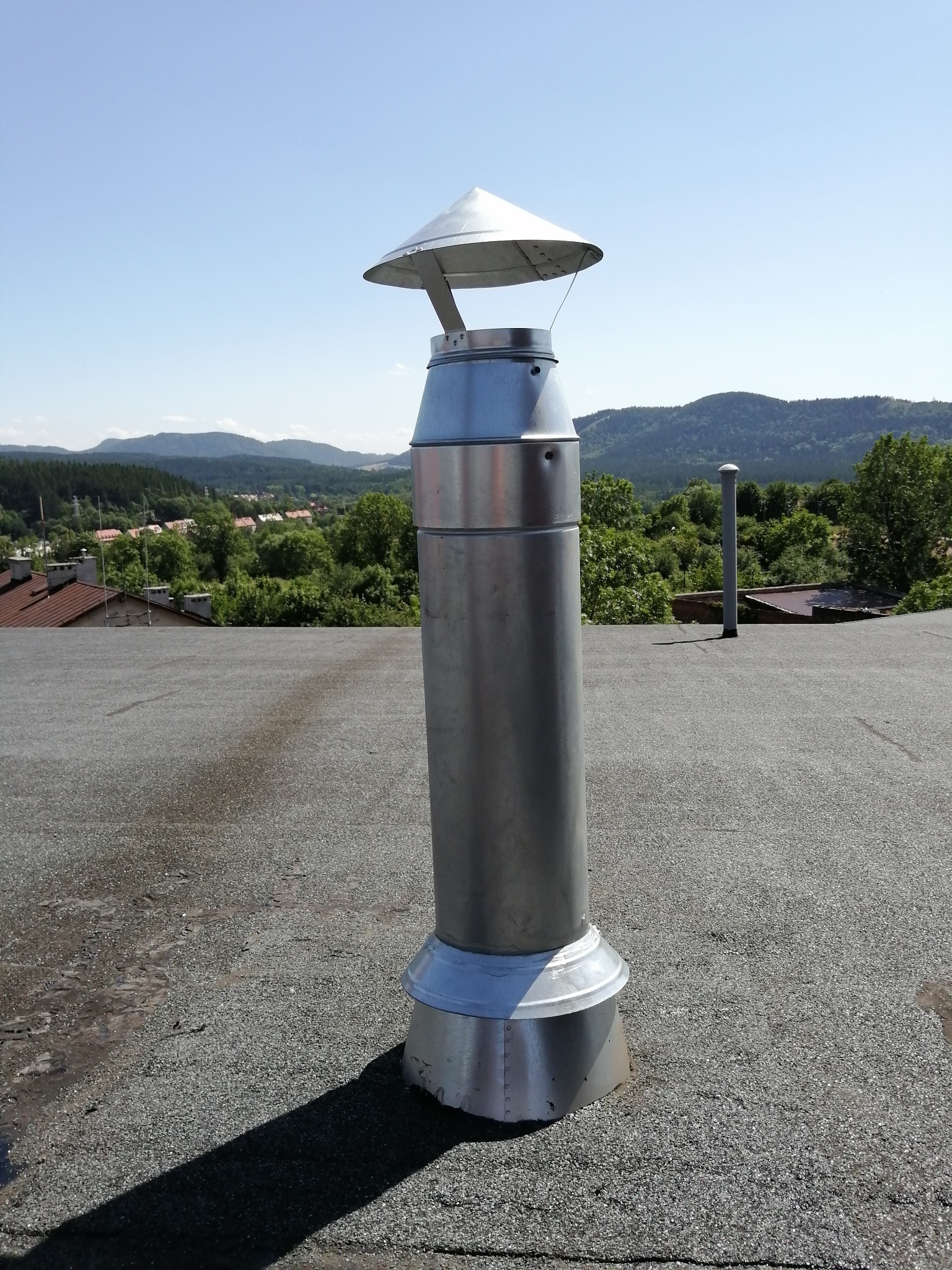 Metalowy komin wentylacyjny z daszkiem na płaskim dachu, w tle zalesione wzgórza pod błękitnym niebem.