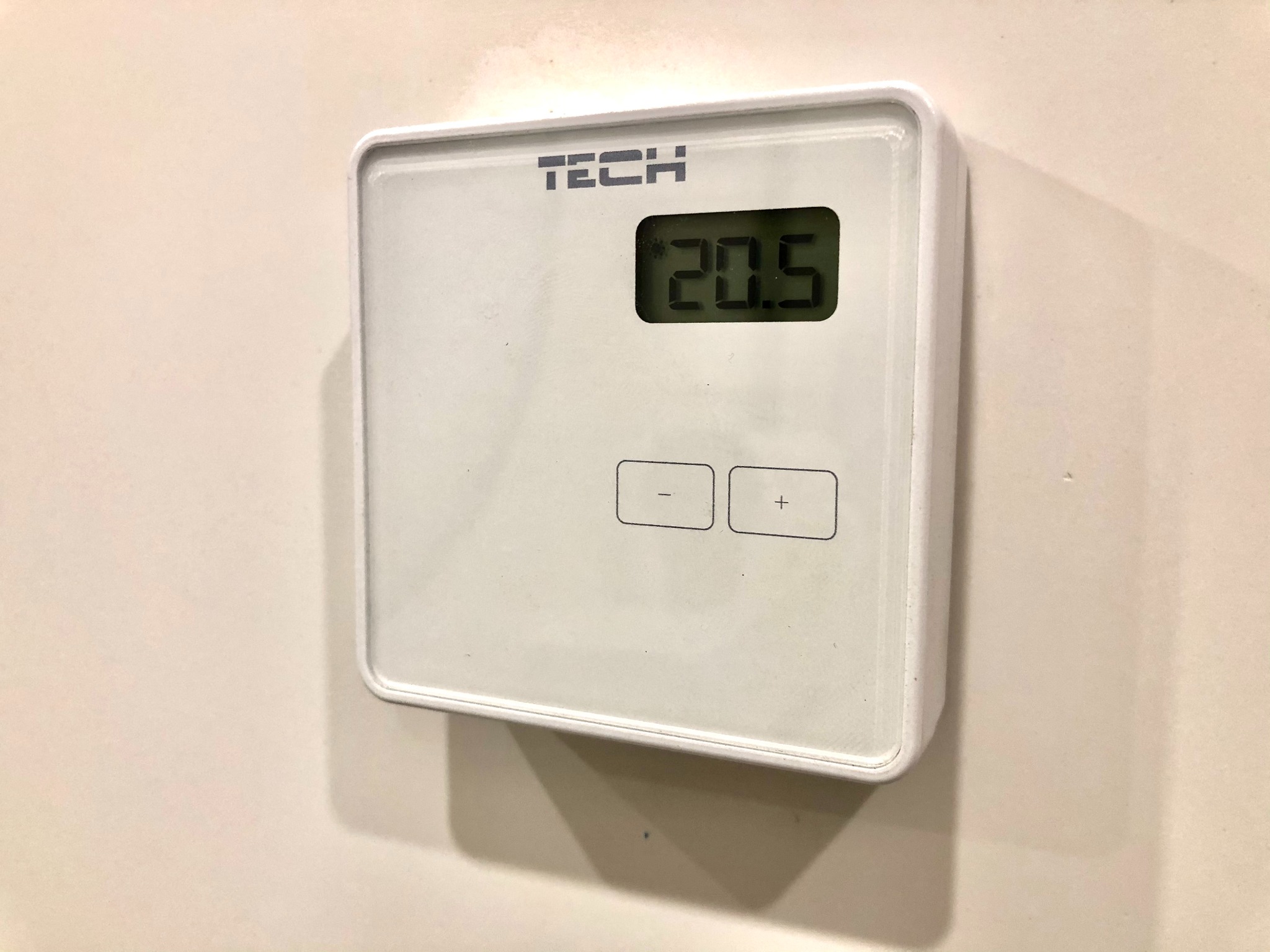 Biały, kwadratowy regulator temperatury TECH zamontowany na jasnej ścianie, wyświetlający temperaturę 20.5 stopni Celsjusza, z widocznymi przyciskami plus i minus.