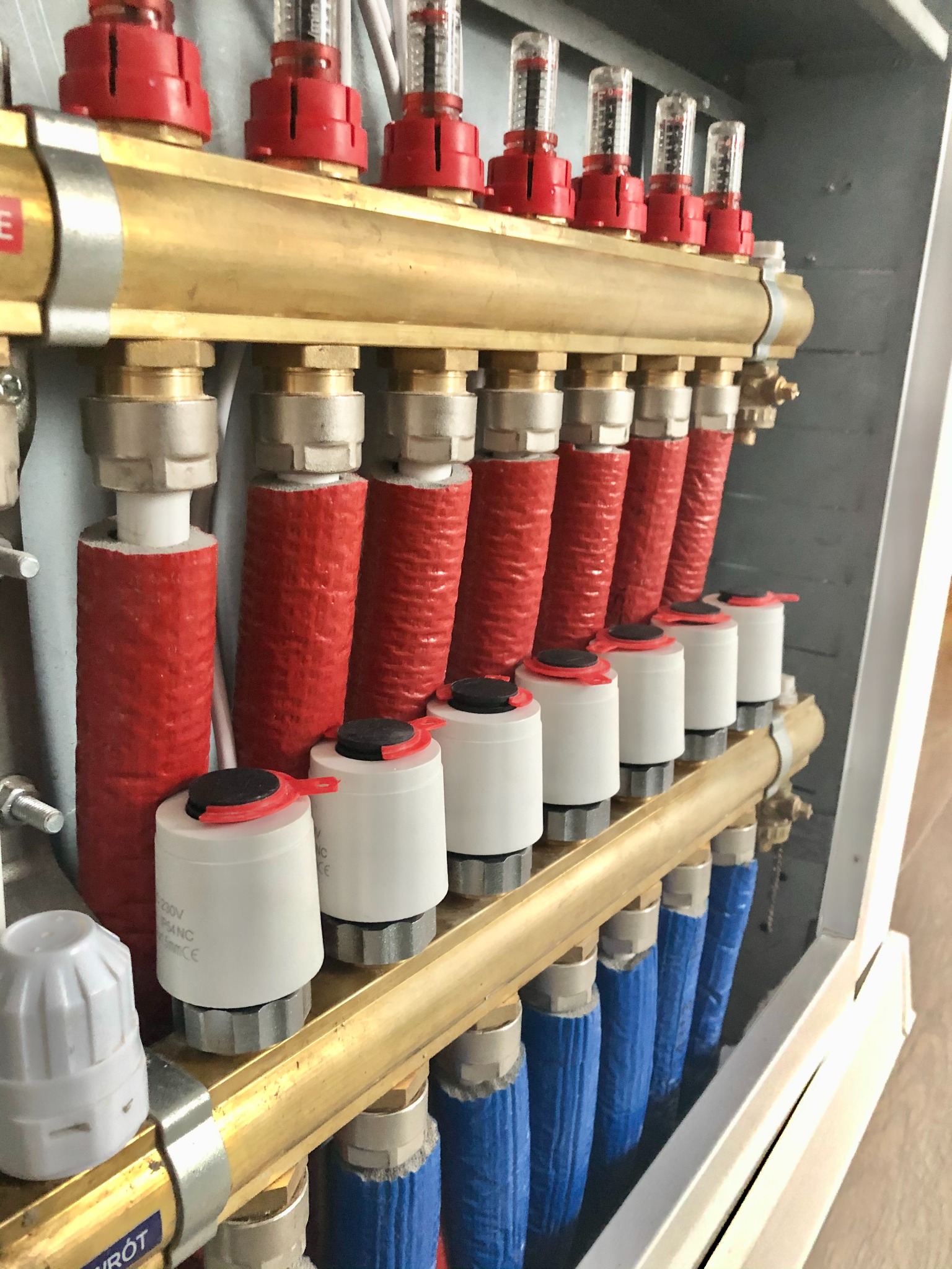 Rozdzielacz ogrzewania podłogowego z widocznymi przepływomierzami, zaworami termostatycznymi i izolowanymi rurami w kolorach czerwonym i niebieskim, zamontowany w skrzynce.