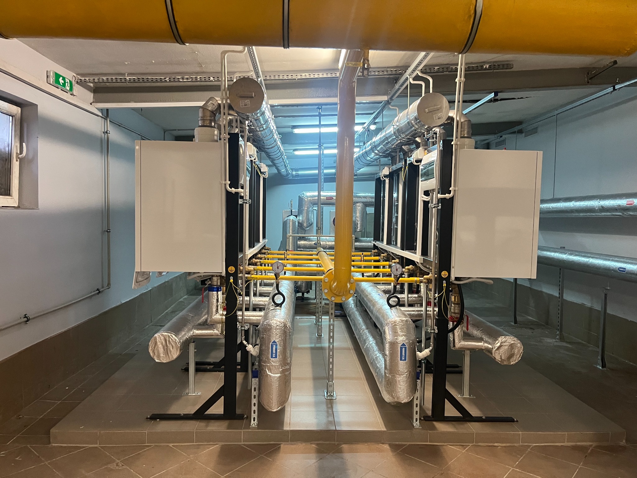 Kompleksowa instalacja grzewcza z rurami w izolacji i żółtymi elementami, widok od frontu. W tle inne elementy instalacji w pomieszczeniu technicznym.