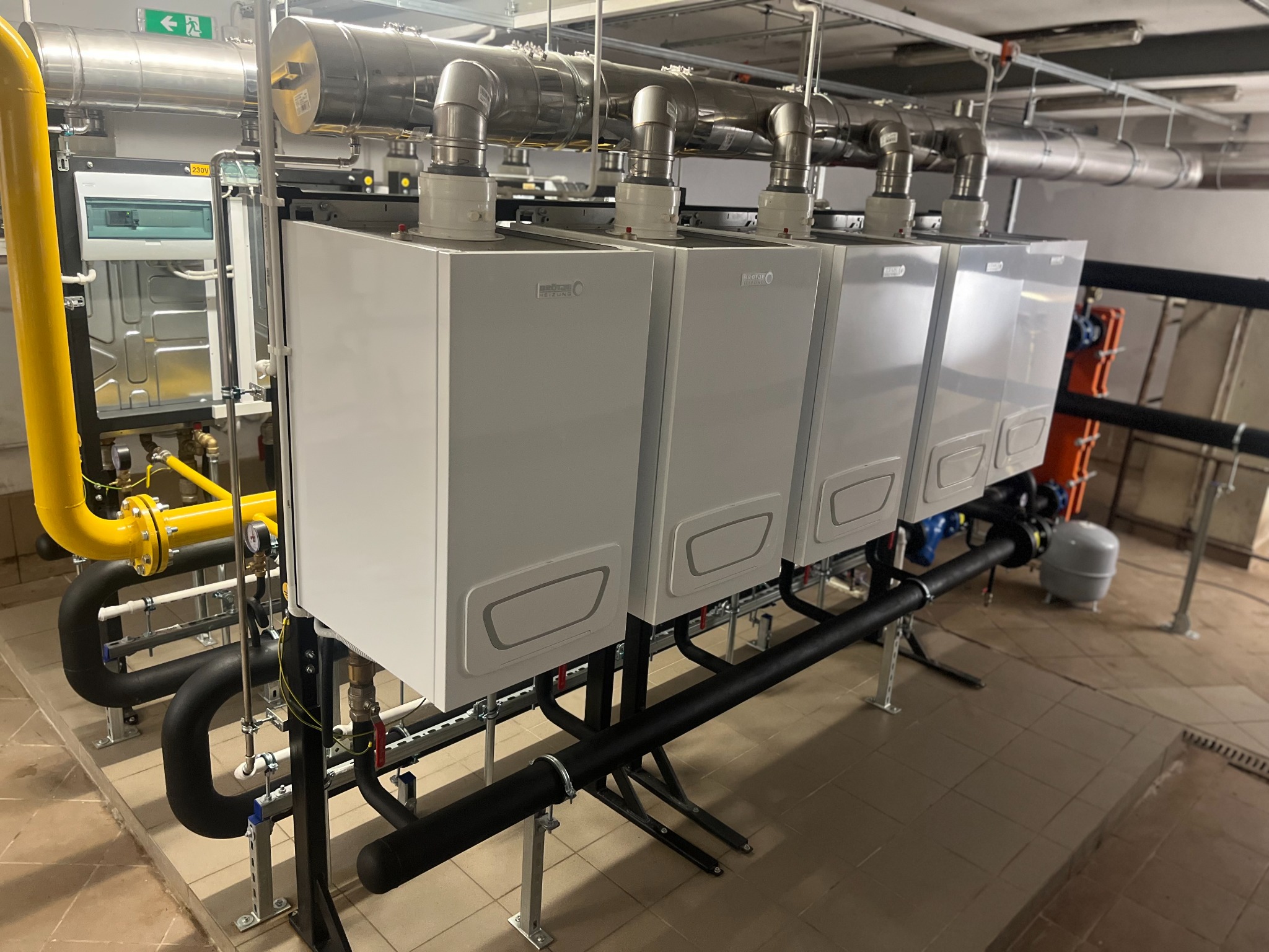 Kaskada czterech nowoczesnych, białych kotłów grzewczych Beretta, z widocznymi rurami i instalacją. Żółta rura gazowa po lewej stronie kadru.