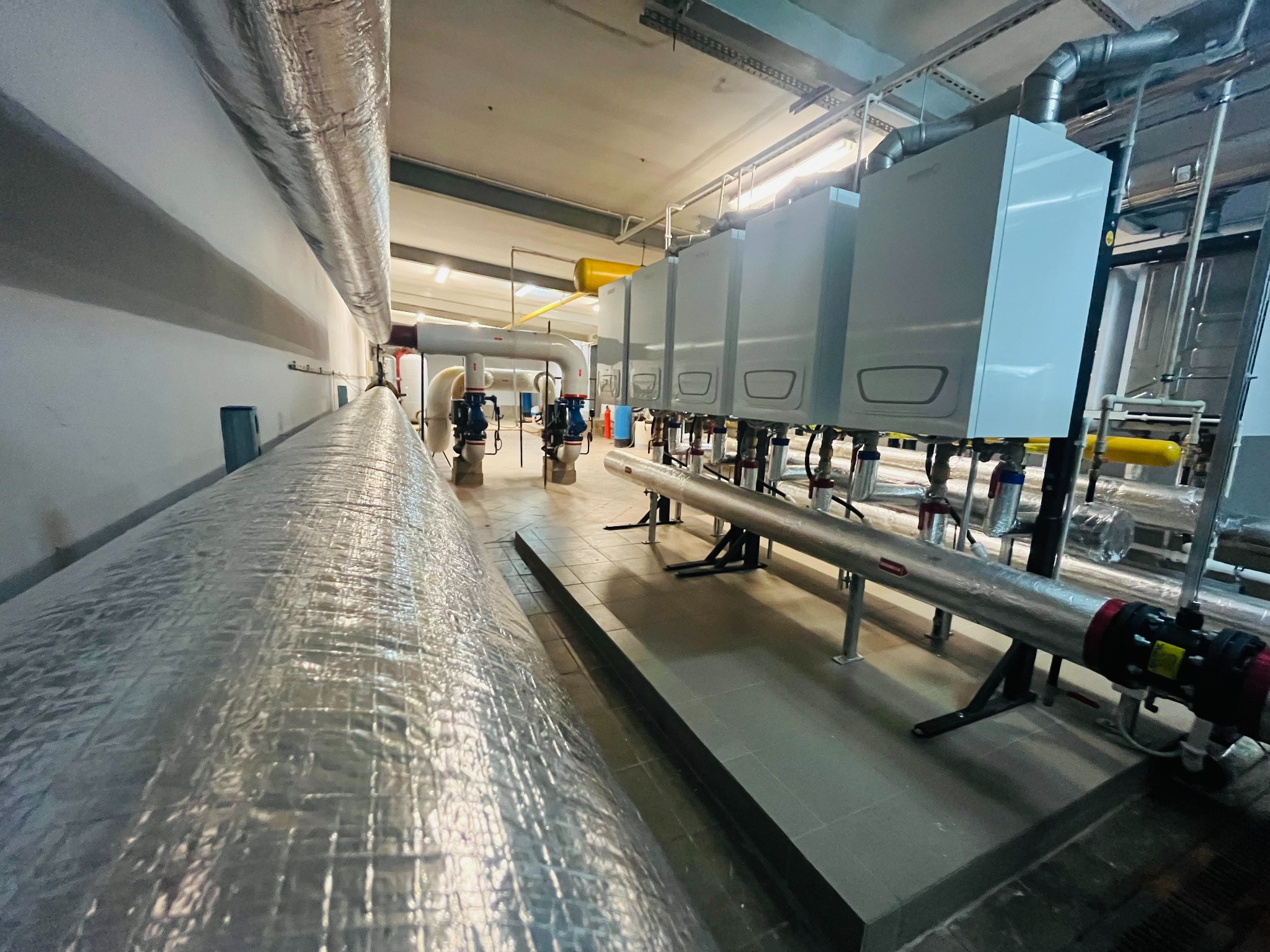 Rząd białych kotłów grzewczych na ścianie, z widocznymi rurami i instalacją, w pomieszczeniu technicznym z oświetleniem i izolowanymi rurami.
