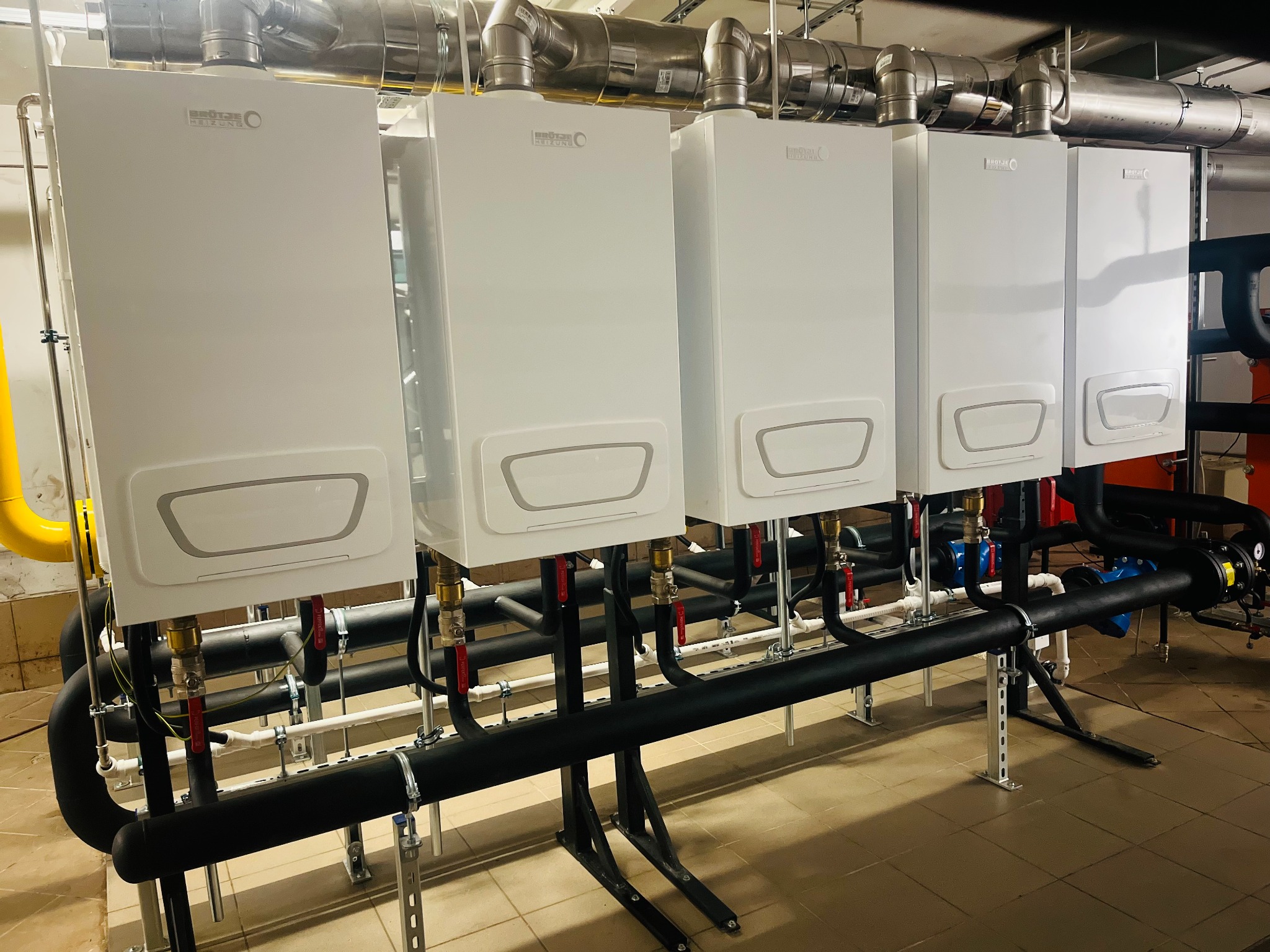Rząd czterech białych kotłów grzewczych Brötje z rurami i zaworami, zamontowanych w kotłowni, widok z dołu. Instalacja w piwnicy.