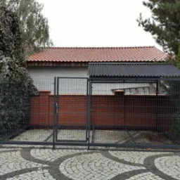 Metalowa konstrukcja z siatki z zadaszeniem, prawdopodobnie kojec dla psa, na tle ceglanego muru i zieleni. Widoczny fragment posesji z kostki brukowej.