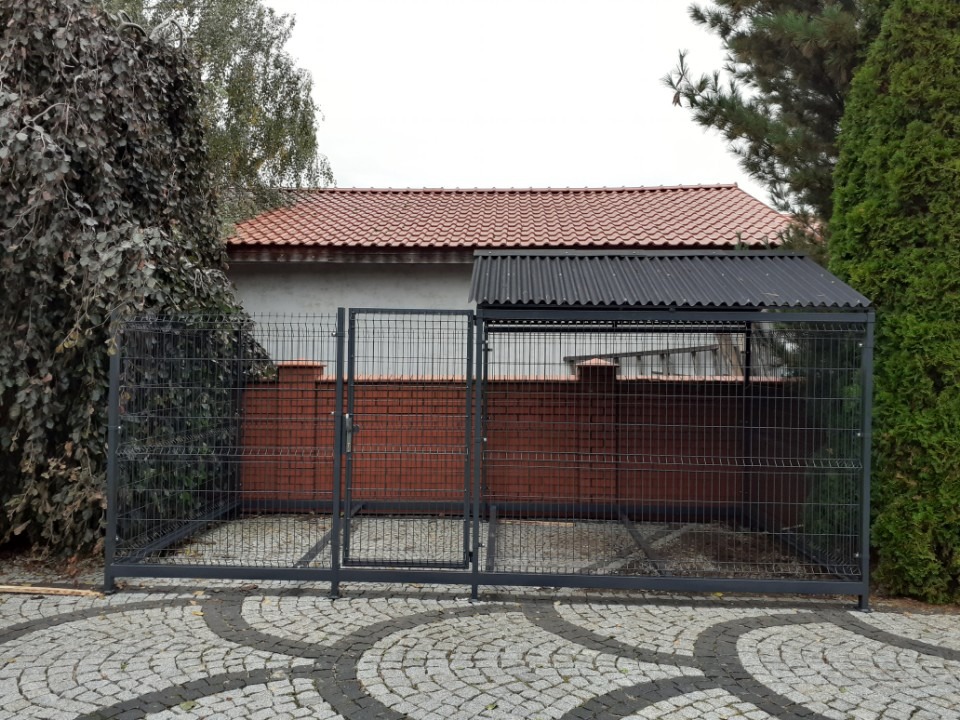 Metalowa konstrukcja z siatki z zadaszeniem, prawdopodobnie kojec dla psa, na tle ceglanego muru i zieleni. Widoczny fragment posesji z kostki brukowej.