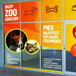 PRZEDSIĘBIORSTWO HANDLOWO-USŁUGOWE KRZYSZTOF GINAŁ - Okna sklepu zoologicznego z pomarańczowo-żółtymi grafikami psów, kotów, papug, kości i misek, oraz napisy informacyjne o godzinach otwarcia i asortymencie.