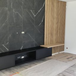 SZYMAŁA TARSYCJUSZ - Nowoczesna szafka RTV w kolorze czarnym, zintegrowana z drewnianą lamelą i półką na tle ściany z płytek imitujących marmur. Minimalistyczny design, widoczne gniazdka.