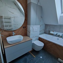 SZYMAŁA TARSYCJUSZ - Nowoczesna łazienka na poddaszu z okrągłym lustrem, umywalką na szafce, toaletą i wanną. Biało-brązowe płytki imitujące marmur i drewno. Ukończony projekt.