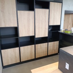 SZYMAŁA TARSYCJUSZ - Nowoczesna szafa wnękowa z drewnianymi frontami i czarnymi ramami, z otwartymi półkami i szufladami, w jasnym wnętrzu.