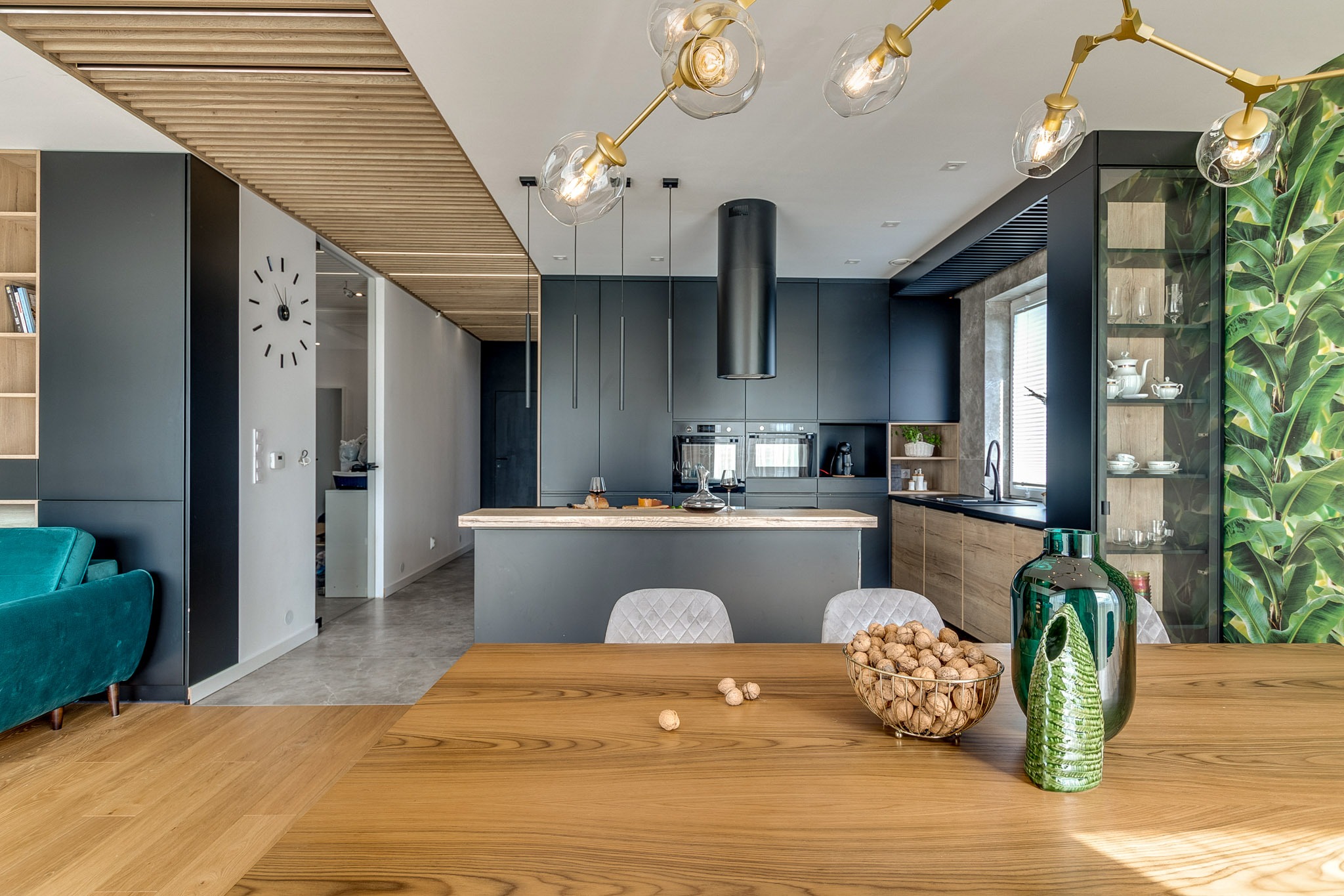 Nowoczesna kuchnia z wyspą i drewnianym stołem. Czarna zabudowa, złote lampy i akcenty zieleni. Na stole dekoracje i owoce. Widok na salon z turkusową sofą.