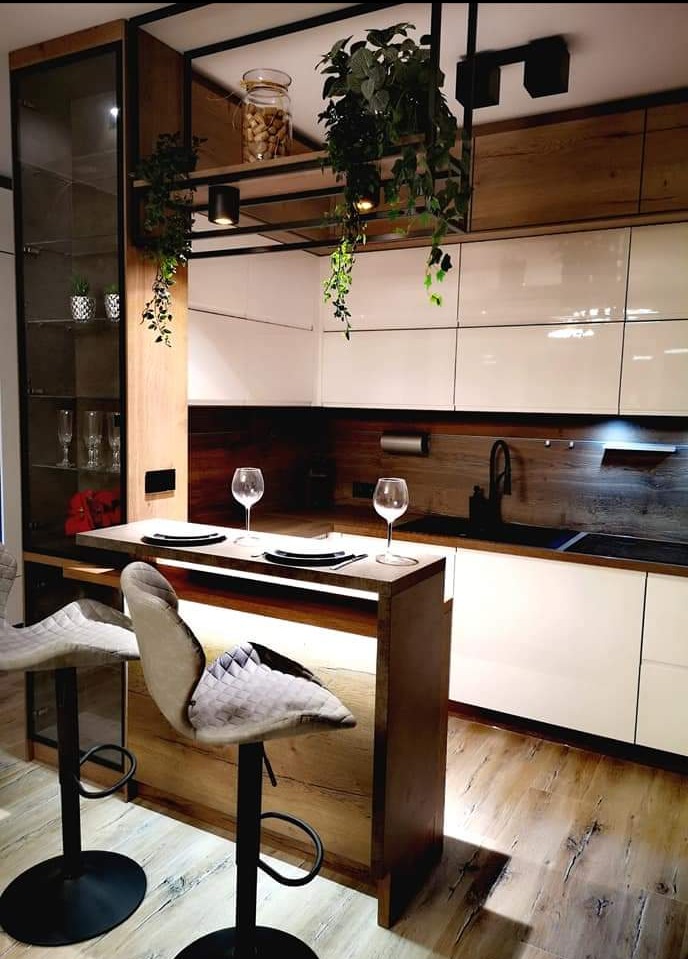 Nowoczesna kuchnia z wyspą i hokerami, białe szafki, drewniane akcenty, oświetlenie punktowe i wiszące rośliny. Nakryty stół z kieliszkami.