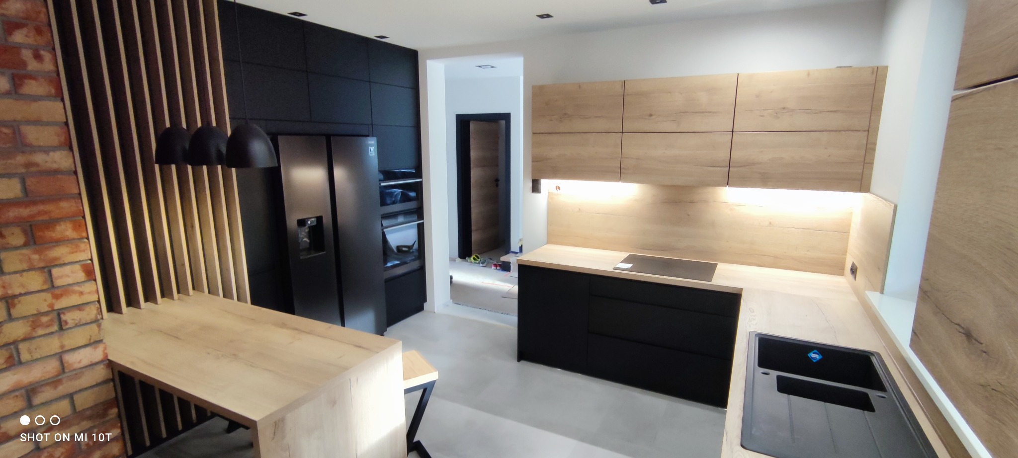 Nowoczesna kuchnia z czarnymi frontami i drewnianymi akcentami, oświetleniem LED i ceglaną ścianą. Widoczny stół z drewnianym blatem i czarnymi nogami.