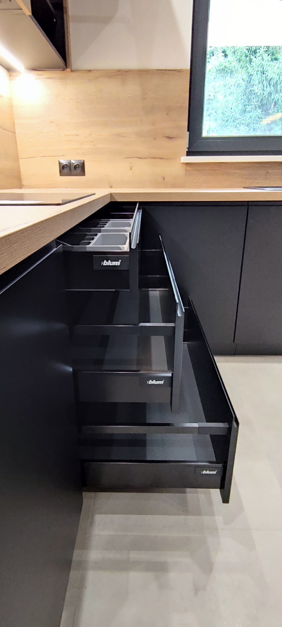 Ciemna, nowoczesna kuchnia z wysuwanymi szufladami Blum, widoczne wewnętrzne przegródki i systemy organizacji przestrzeni. Drewniany blat i okno w tle.