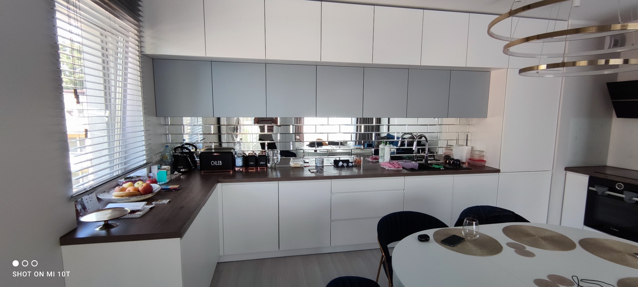 Nowoczesna kuchnia z białymi szafkami, szarymi górnymi szafkami i lustrzanymi płytkami nad blatem. Na blacie akcesoria kuchenne i owoce. Okno z żaluzjami.