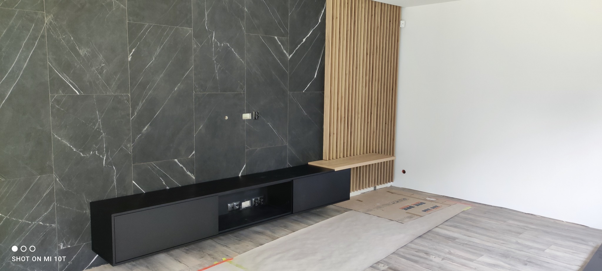 Nowoczesna szafka RTV w kolorze czarnym, zintegrowana z drewnianą lamelą i półką na tle ściany z płytek imitujących marmur. Minimalistyczny design, widoczne gniazdka.