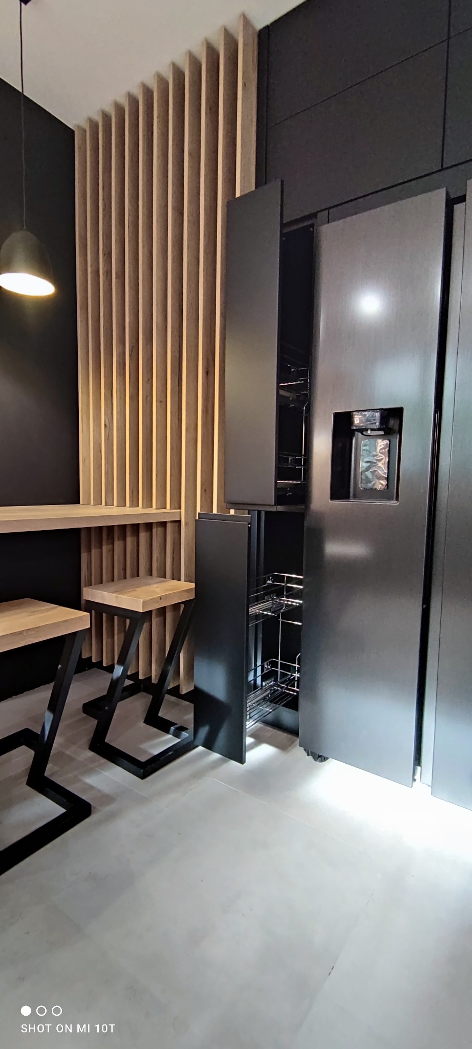 Nowoczesna kuchnia z czarnymi frontami, drewnianymi lamelami i wysuwaną szafką cargo. Minimalistyczny design z oświetleniem punktowym i hokerami.