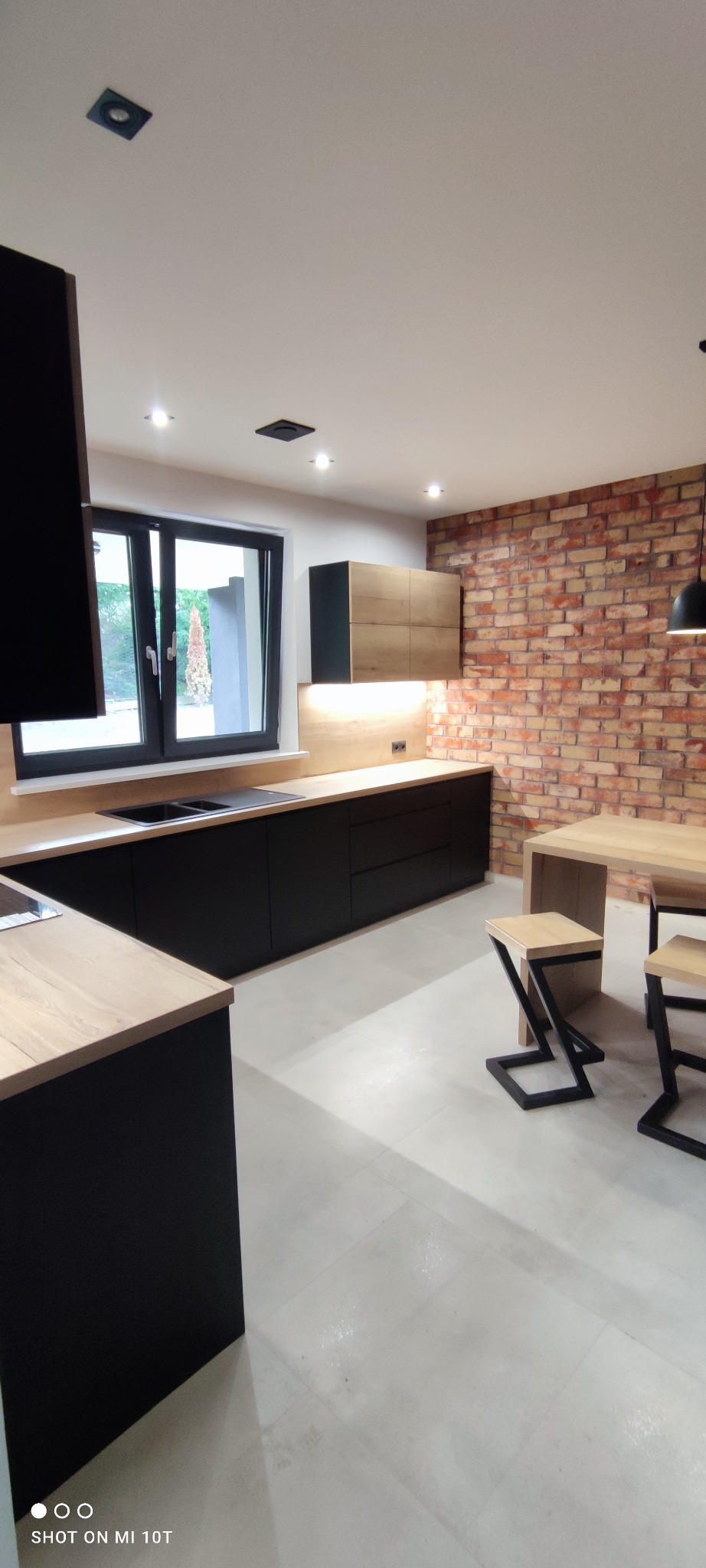 Nowoczesna kuchnia z czarnymi frontami i drewnianym blatem, ceglana ściana w tle, stół z krzesłami o geometrycznych nogach. Minimalistyczny design, jasne wnętrze.