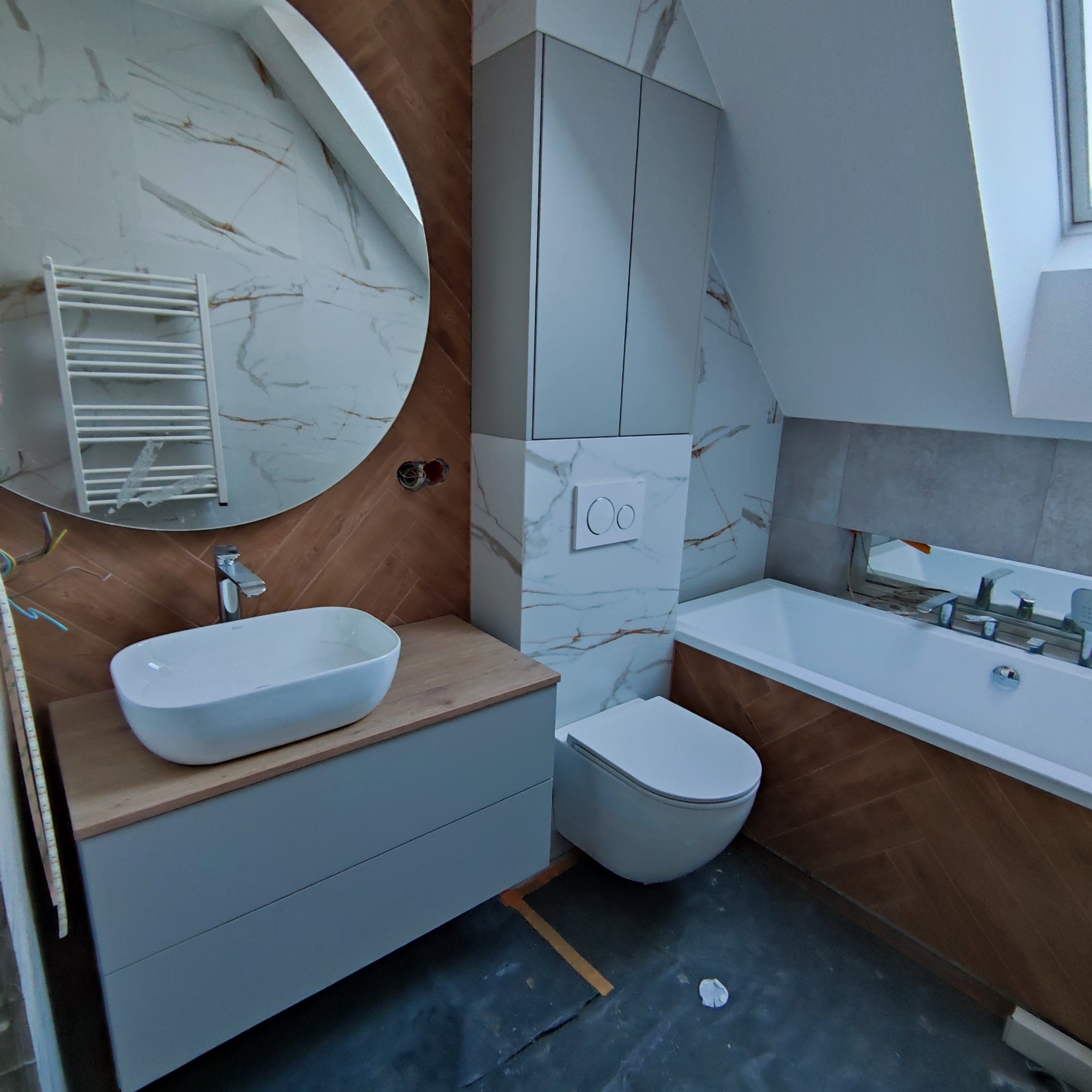 Nowoczesna łazienka na poddaszu z okrągłym lustrem, umywalką na szafce, toaletą i wanną. Biało-brązowe płytki imitujące marmur i drewno. Ukończony projekt.