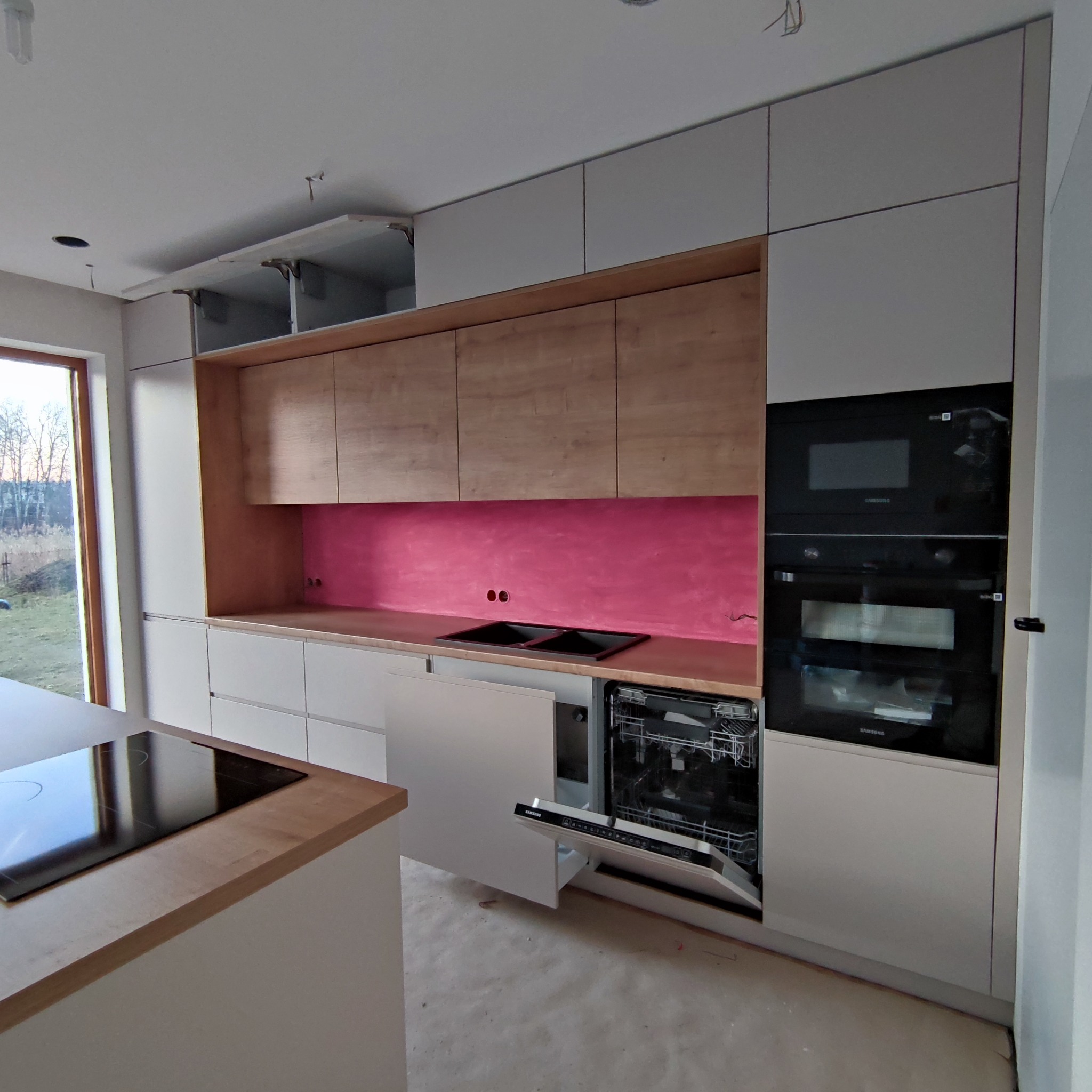 Jasna kuchnia z drewnianymi akcentami i różowym panelem nad blatem. Otwarta zmywarka, piekarnik i mikrofalówka w zabudowie. Widok na zewnątrz przez okno.