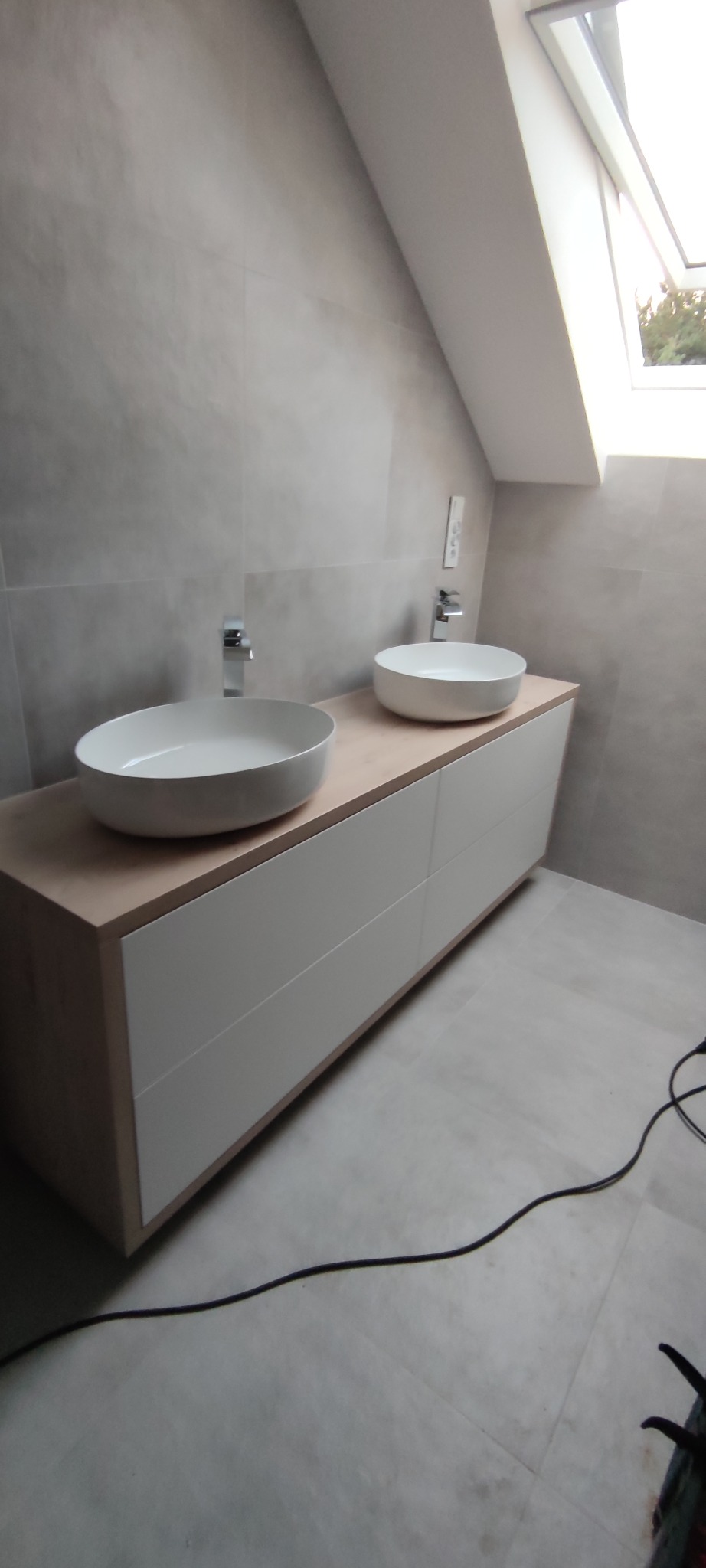 Nowoczesna łazienka z dwiema okrągłymi umywalkami nablatowymi i szafką z białymi frontami, oświetlona światłem z okna dachowego. Minimalistyczny design.