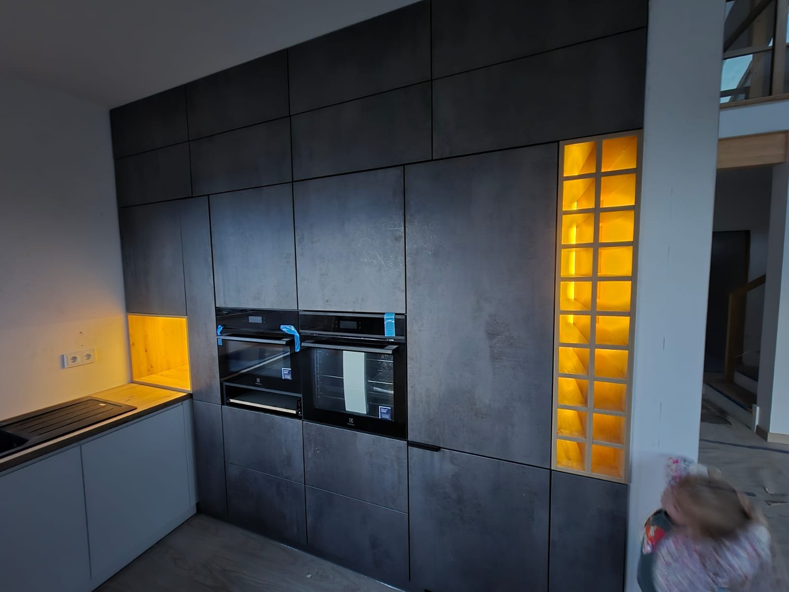 Nowoczesna kuchnia z szarymi frontami, wbudowanymi piekarnikami i podświetlanymi wnękami. Minimalistyczny design z akcentami ciepłego światła.