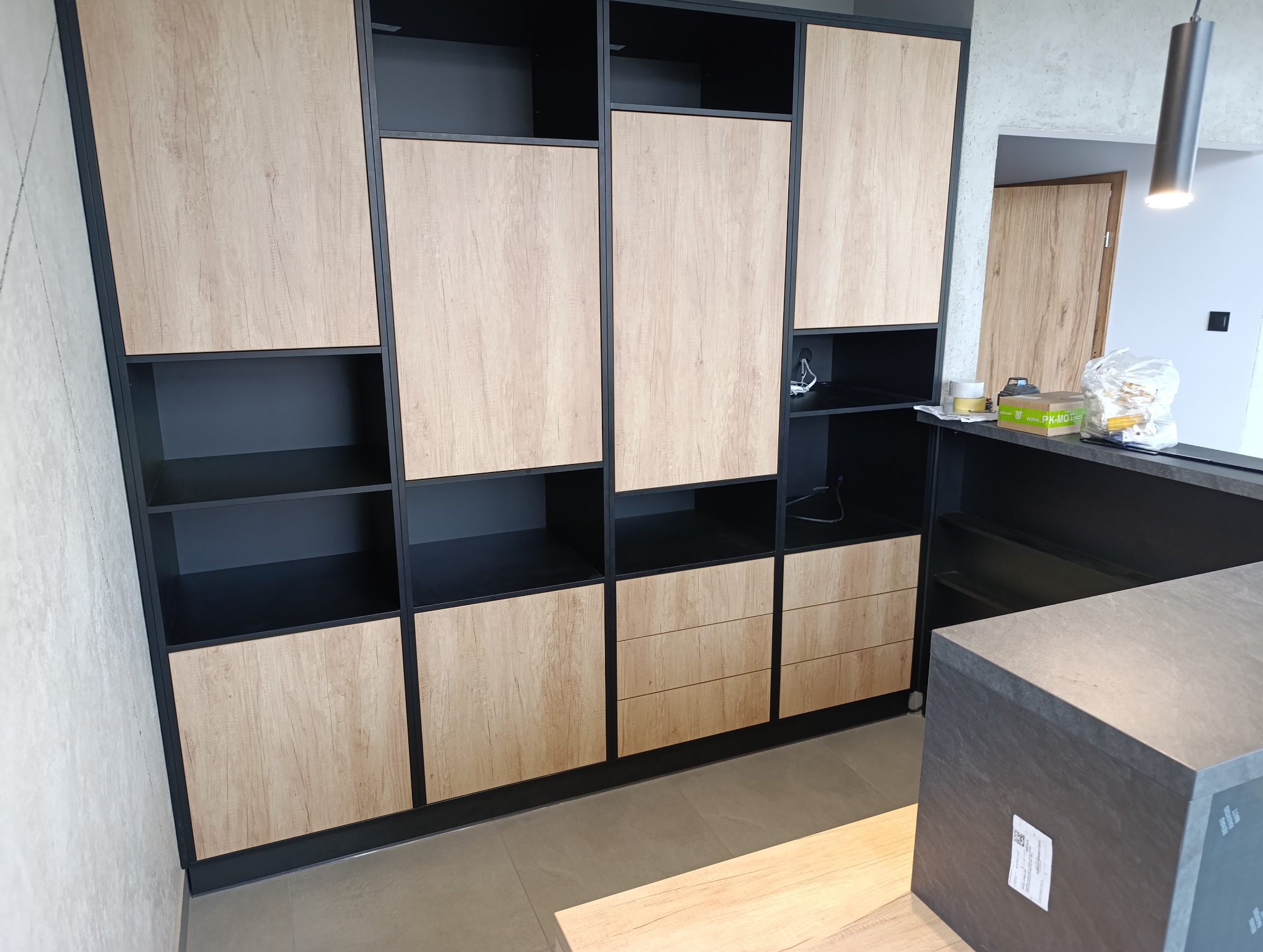 Nowoczesna szafa wnękowa z drewnianymi frontami i czarnymi ramami, z otwartymi półkami i szufladami, w jasnym wnętrzu.