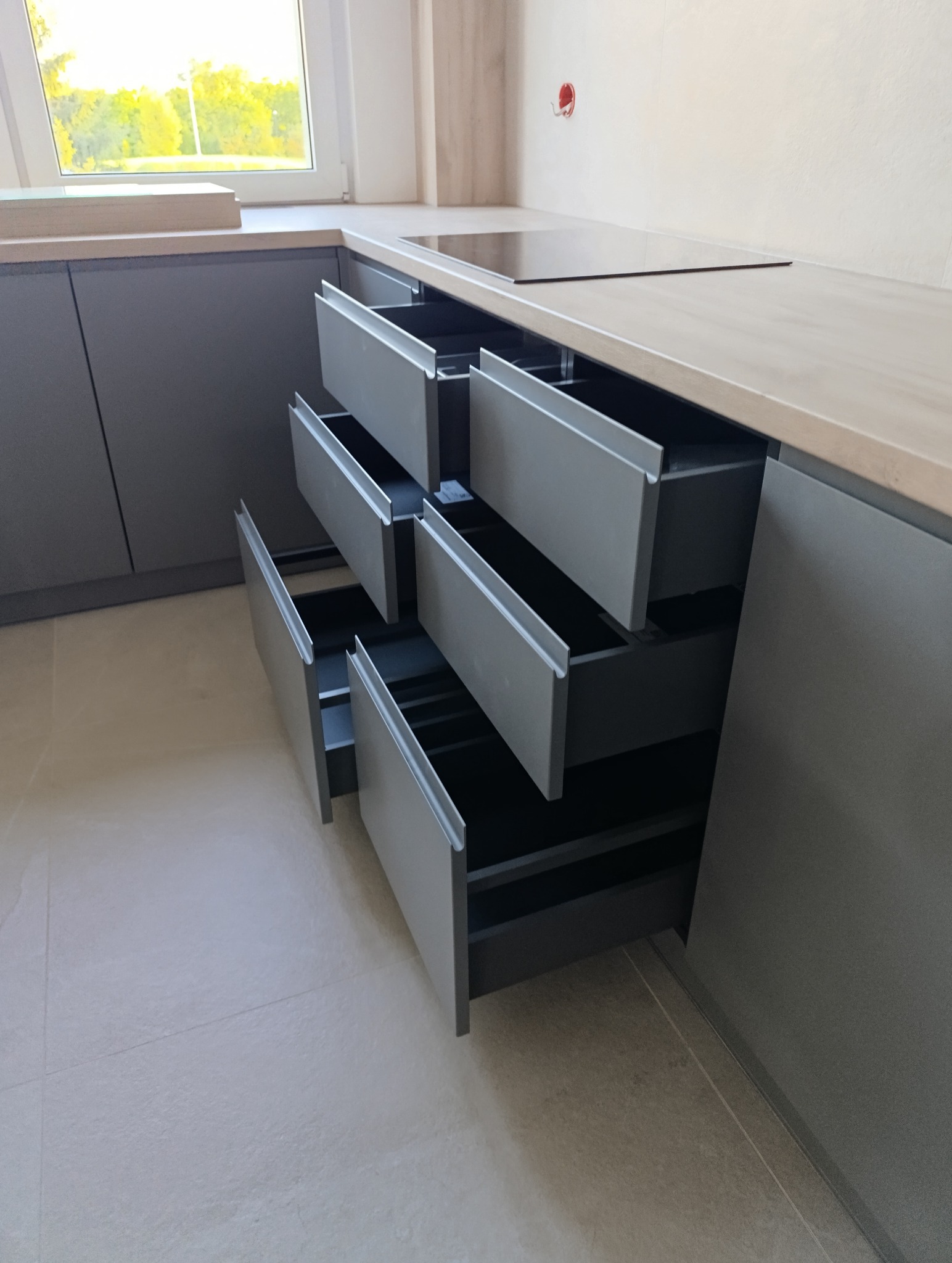 Szare, nowoczesne szafki kuchenne z wysuwanymi szufladami, widok z bliska na otwarte szuflady, jasne płytki podłogowe i blat, okno w tle.